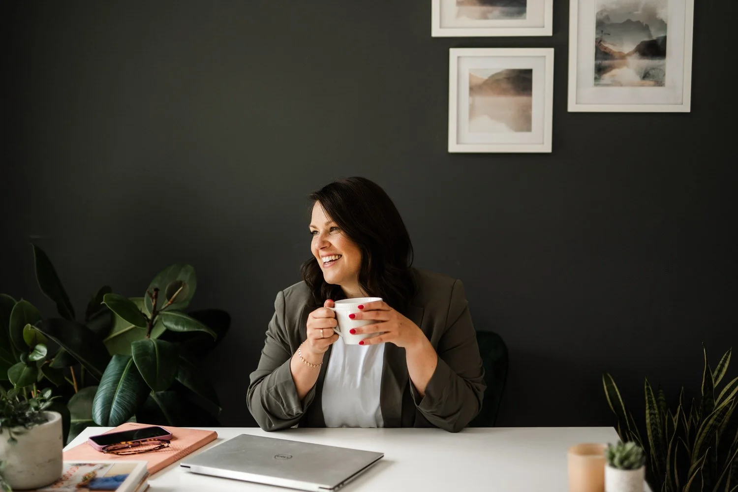 A woman with dark hair smiling while holding a white mug, sitting at a desk with plants, a closed laptop, and notebooks, against a dark wall with framed pictures.