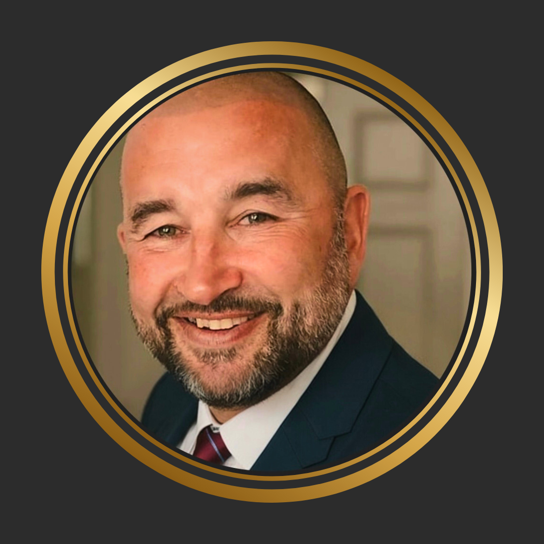 Close-up of a smiling man with a beard, bald head, dressed in a suit and tie, in a professional setting.