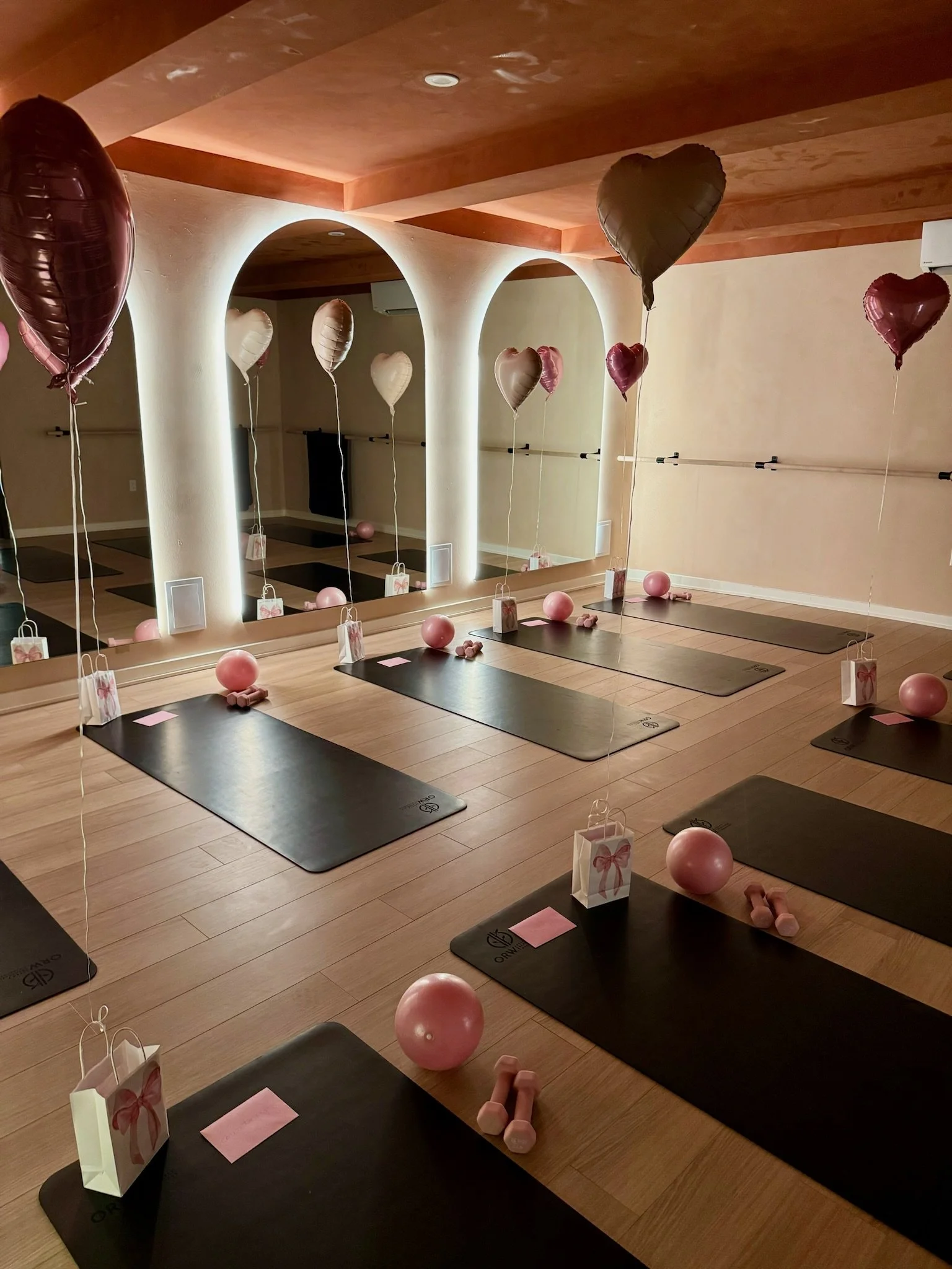 Indoor room decorated with heart-shaped balloons, gift bags, pink balloons, and foam dumbbells on yoga mats, wooden ceiling, mirrors, and light-colored walls.