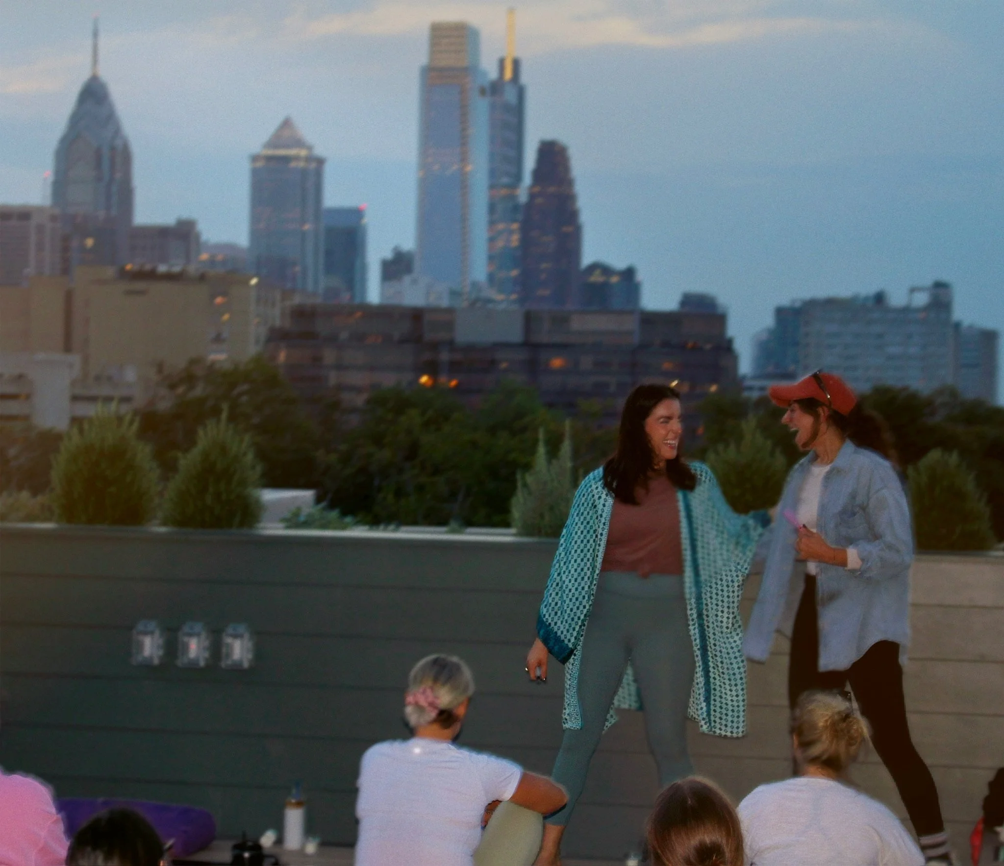 Two women are standing outside, smiling and engaging in conversation, with a cityscape including tall skyscrapers in the background during dusk.