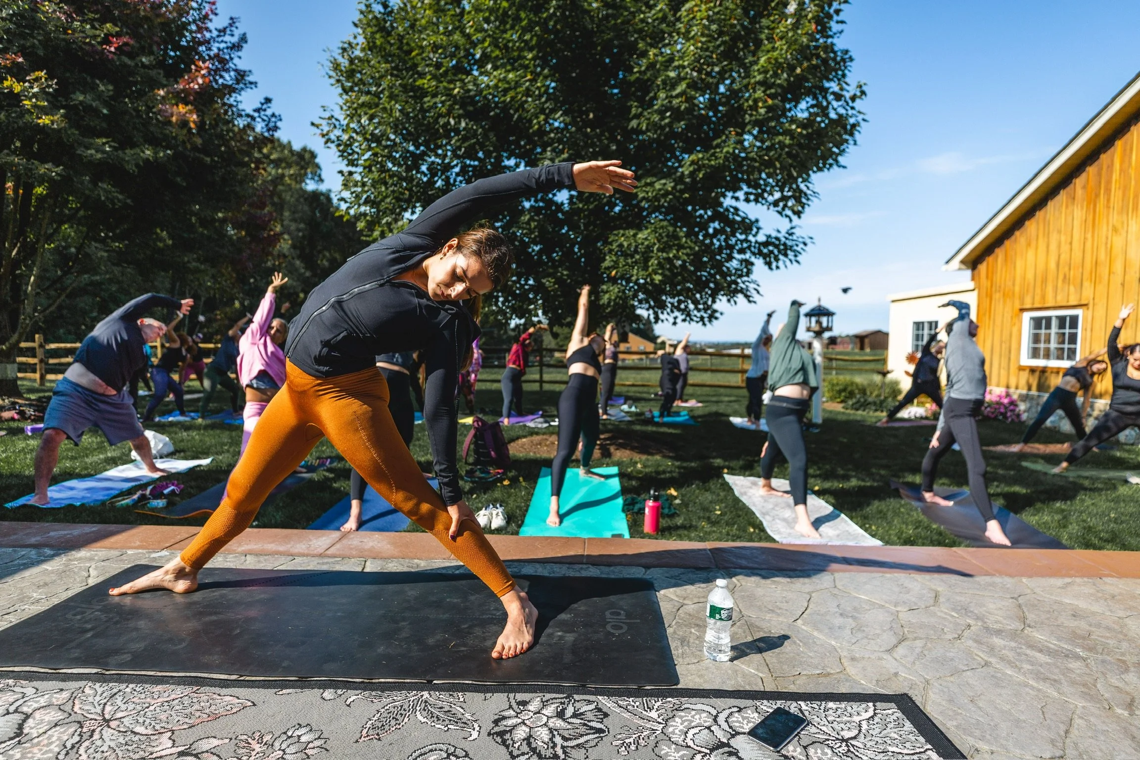 A group of people practicing yoga outdoors in a park on a sunny day, with trees and a yellow building in the background.