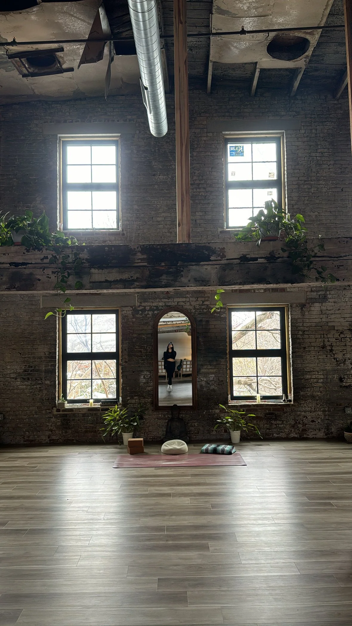 A spacious room with exposed brick walls and large windows letting in natural light. There are plants on the window sills and a mirror on the wall reflecting a person taking a photo. The wooden floor is furnished with a yoga mat, a cushion, and some folded blankets for a yoga or meditation space.