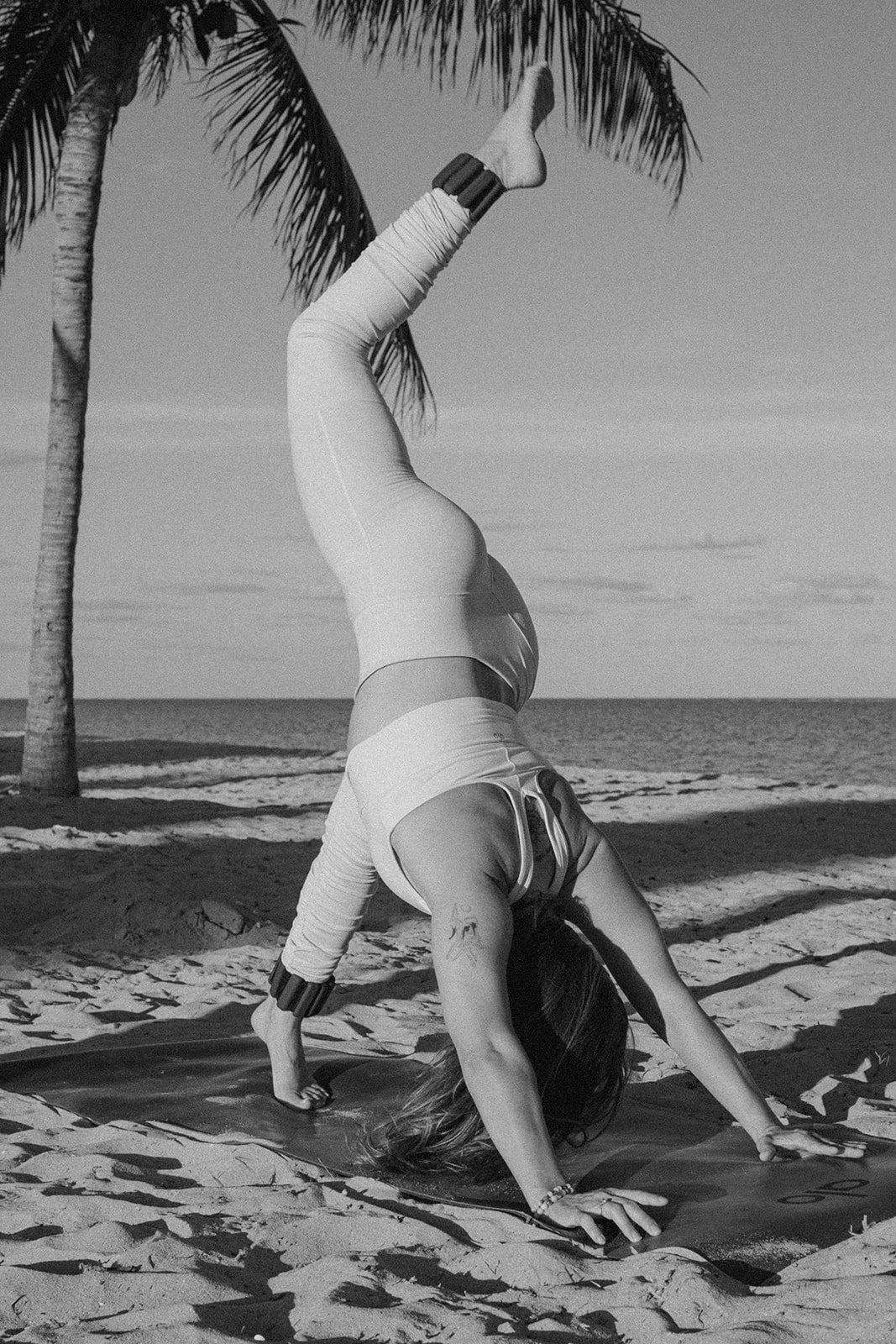 A woman performing a headstand yoga pose on a beach with palm trees and the ocean in the background, in black and white.
