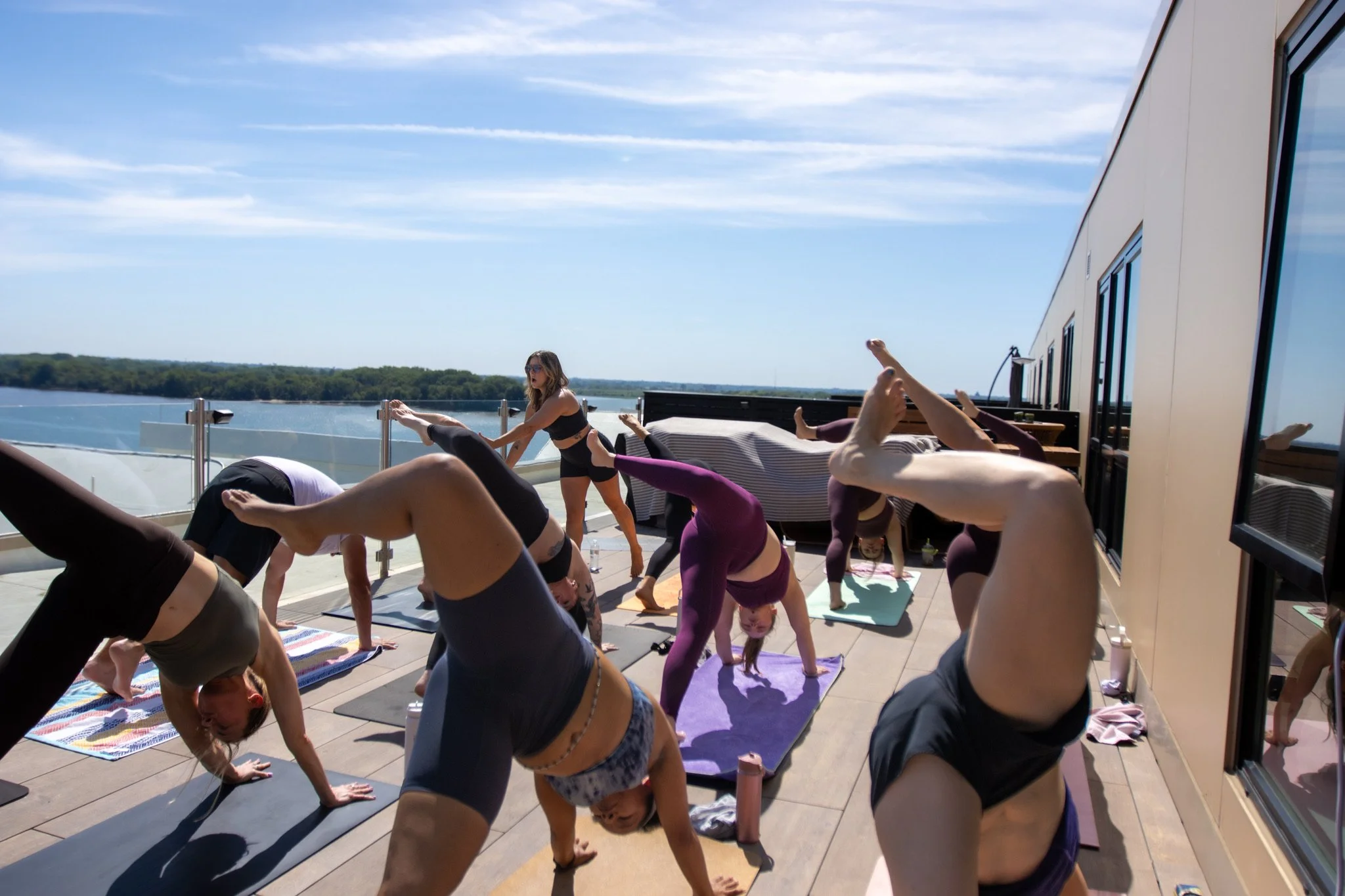A group of people practicing yoga on outdoor mats on a rooftop balcony, with a scenic view of water and trees in the background, during daytime.