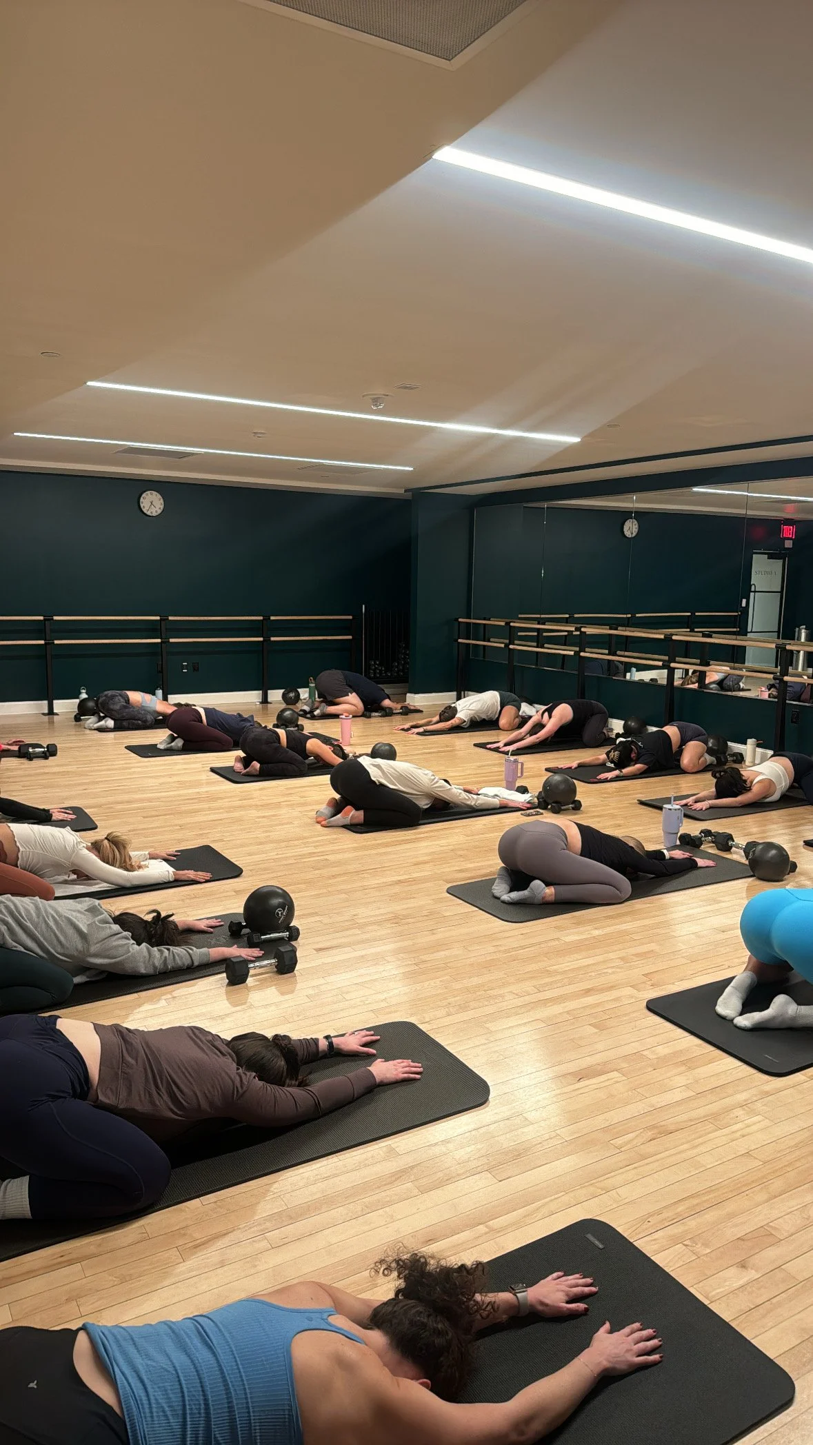 People participating in a yoga class lying on mats in a dance studio with mirrors, a clock, and overhead lighting.