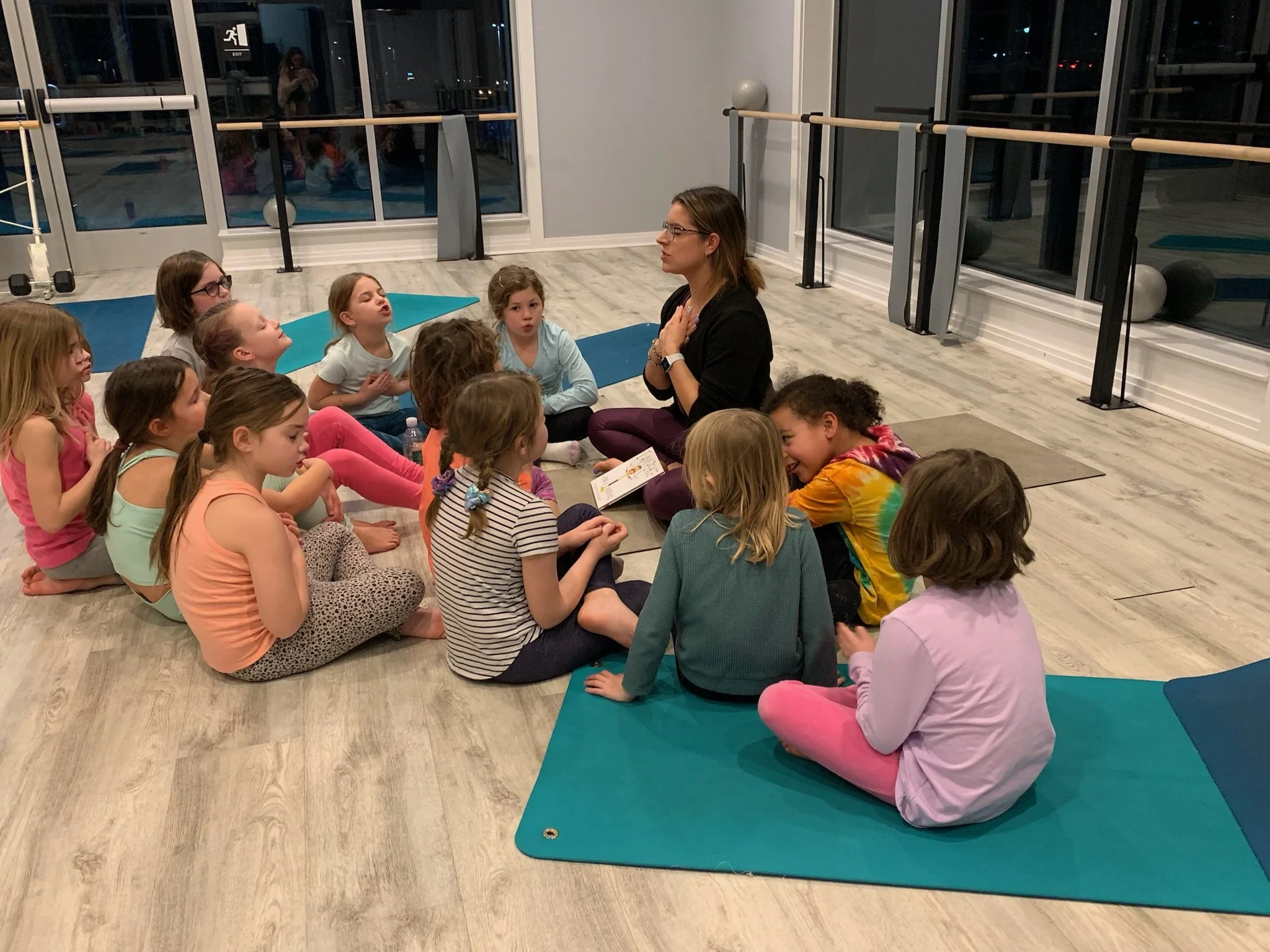 A group of children sitting cross-legged on yoga mats in a studio with a woman leading a meditation or mindfulness session, during nighttime visible through large windows.