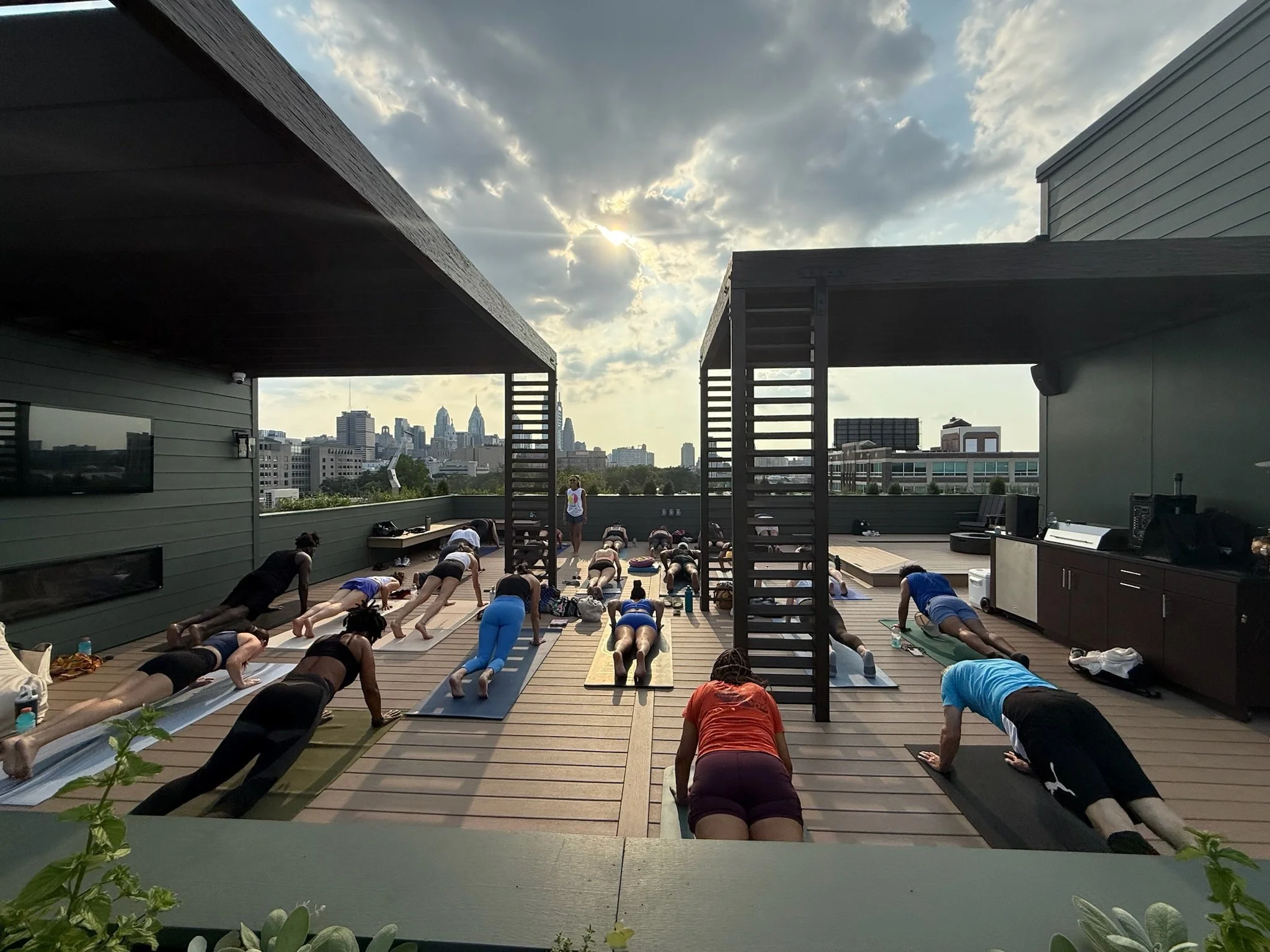 Individuals participating in a rooftop yoga class at sunset, performing downward dog pose on yoga mats with a city skyline in the background.