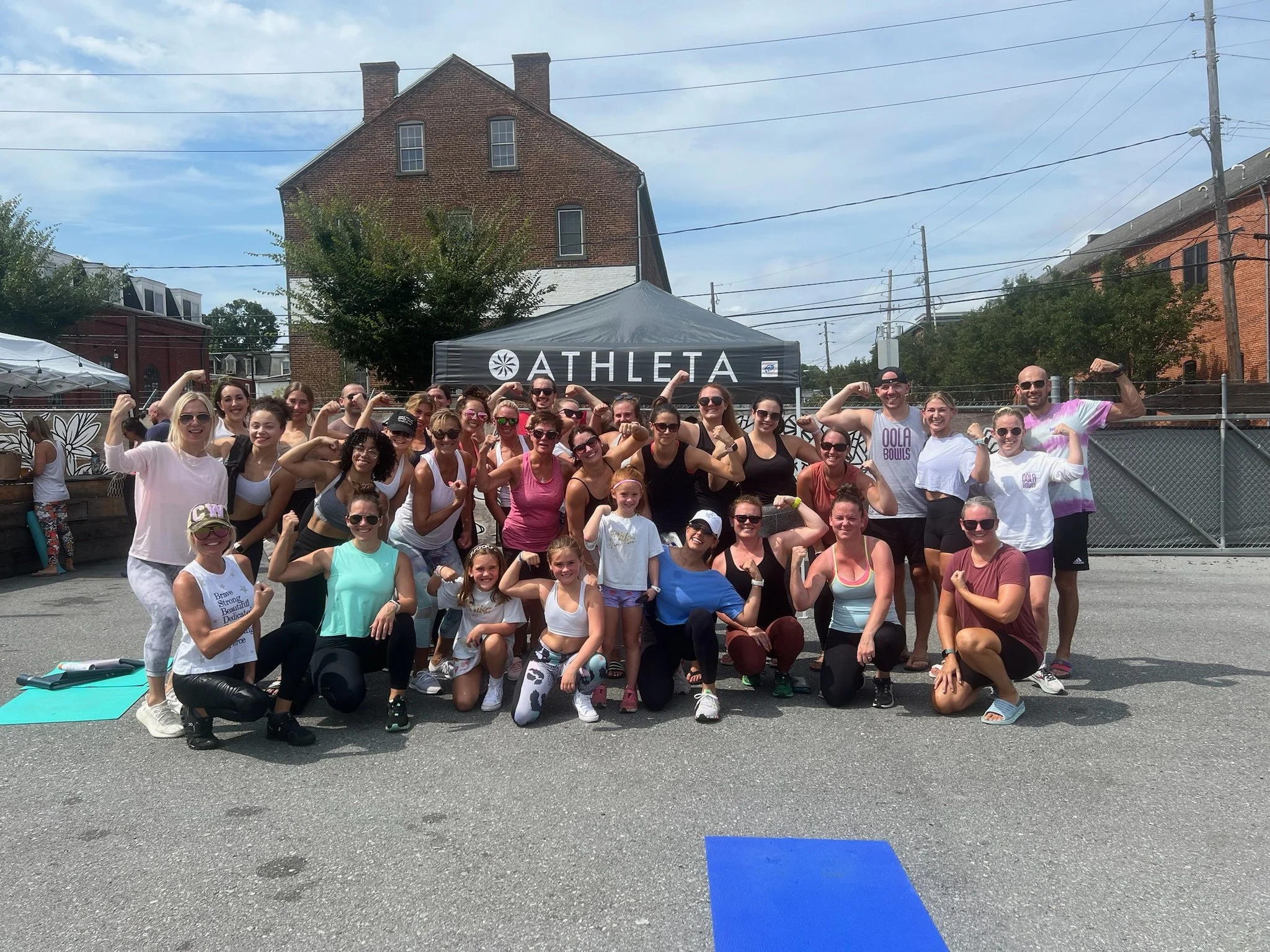Group of people in athletic clothing posing outdoors at a fitness event with a promotional tent that says 'Athleta' in the background.