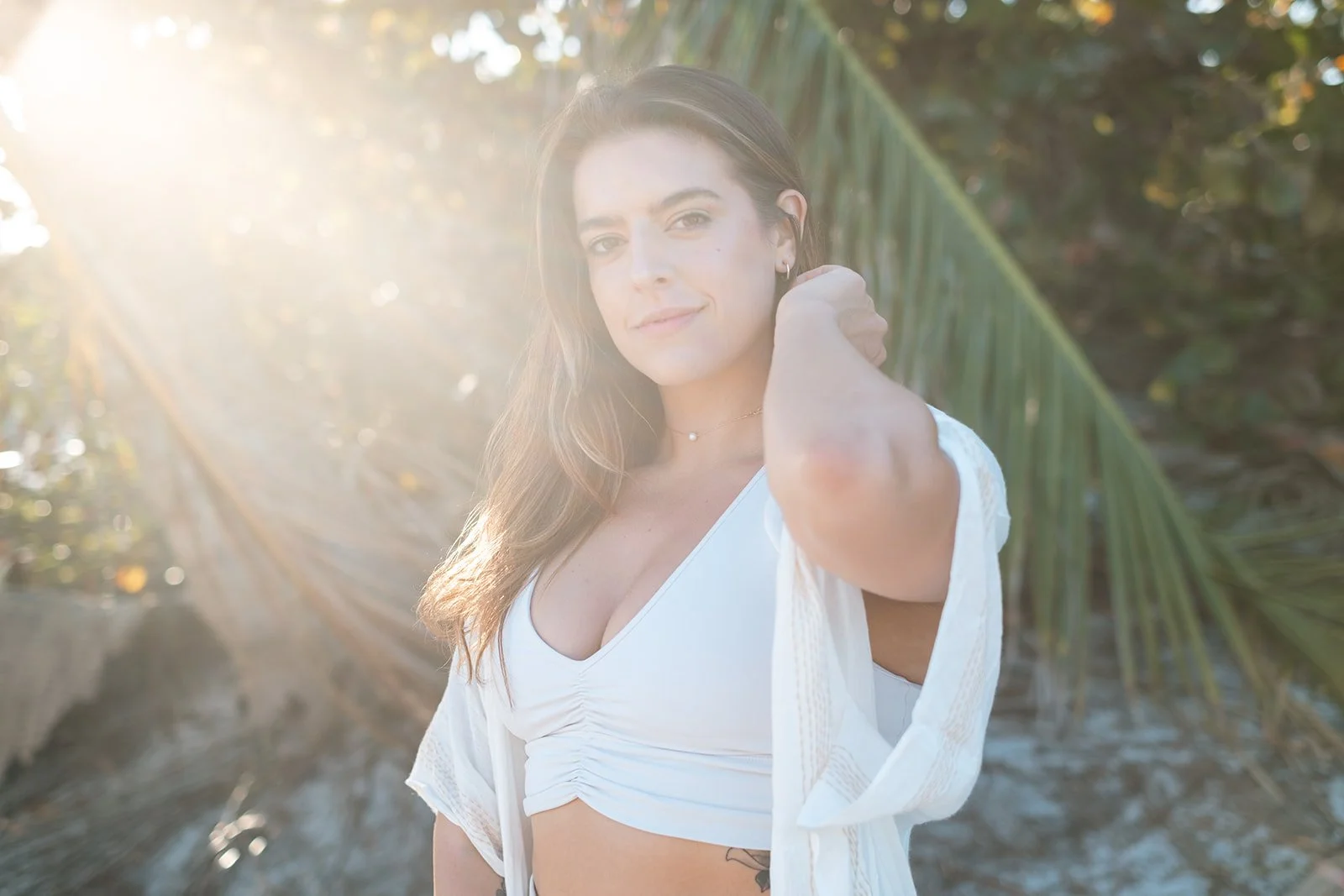 A woman with long brown hair wearing a white top and a light cover-up, standing outdoors near palm trees, with sunlight illuminating her face from the left side.