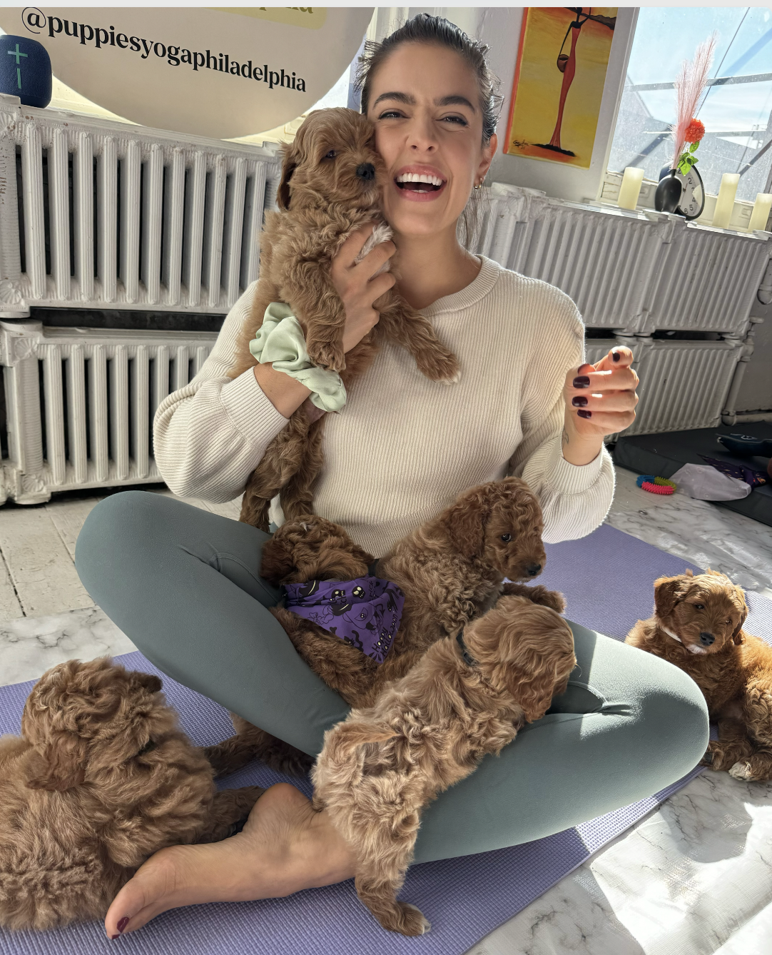 A woman sitting cross-legged on a yoga mat surrounded by several adorable, fluffy brown puppies. She is holding one puppy close to her face, smiling and laughing. The room has a bright window, candles, a clock, and colorful artwork on the walls, creating a cozy atmosphere.