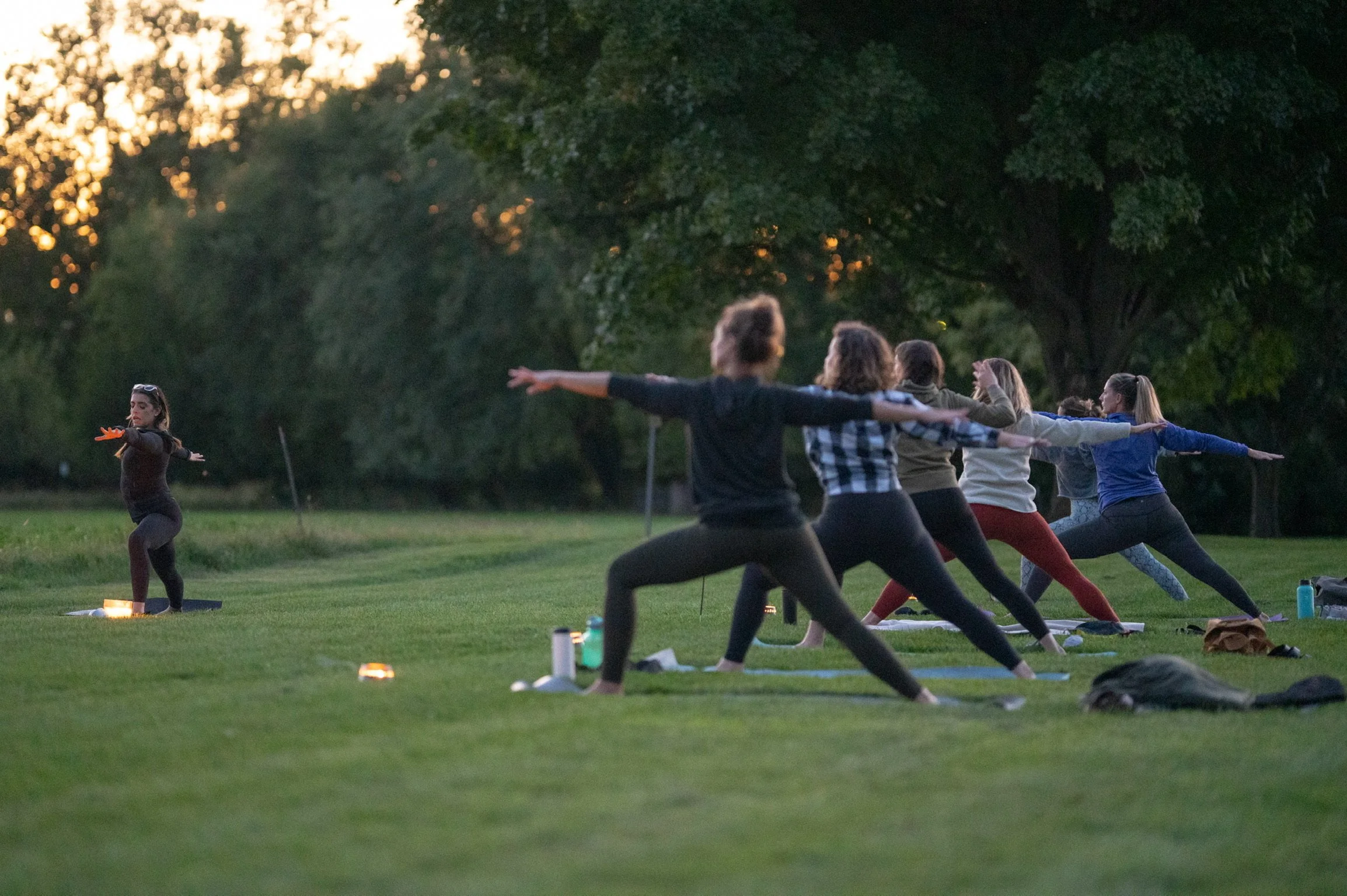 Group of people practicing yoga outdoors at sunset, standing in a lunge pose with arms extended, on a grassy field with trees in the background.