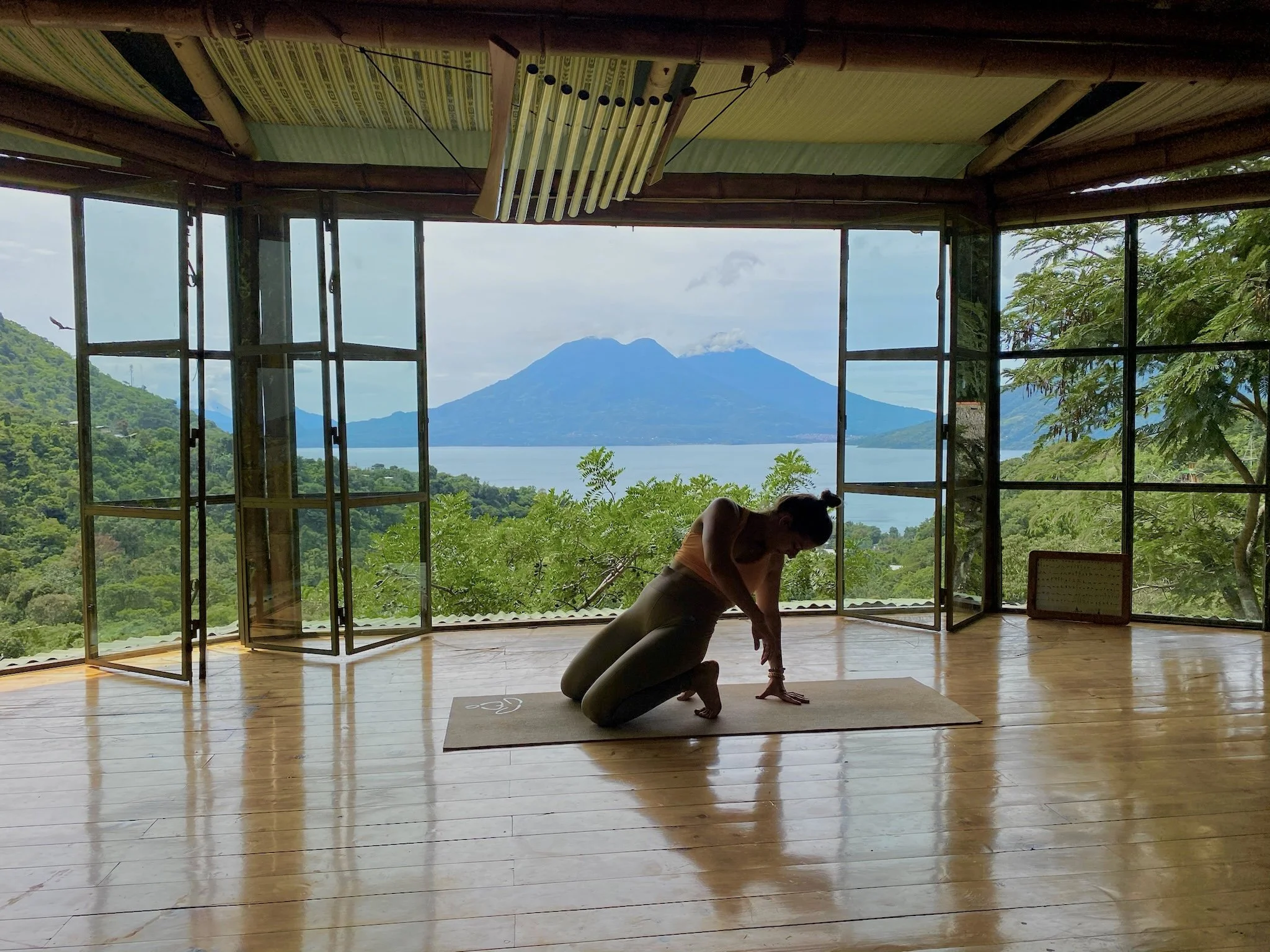 A woman practicing yoga on a mat in a room with open windows, overlooking a scenic view of a lake and mountains in the distance.