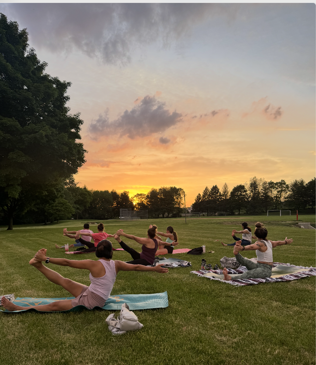 People practicing yoga outdoors on mats during sunset in a park, with trees and sports fields in the background.