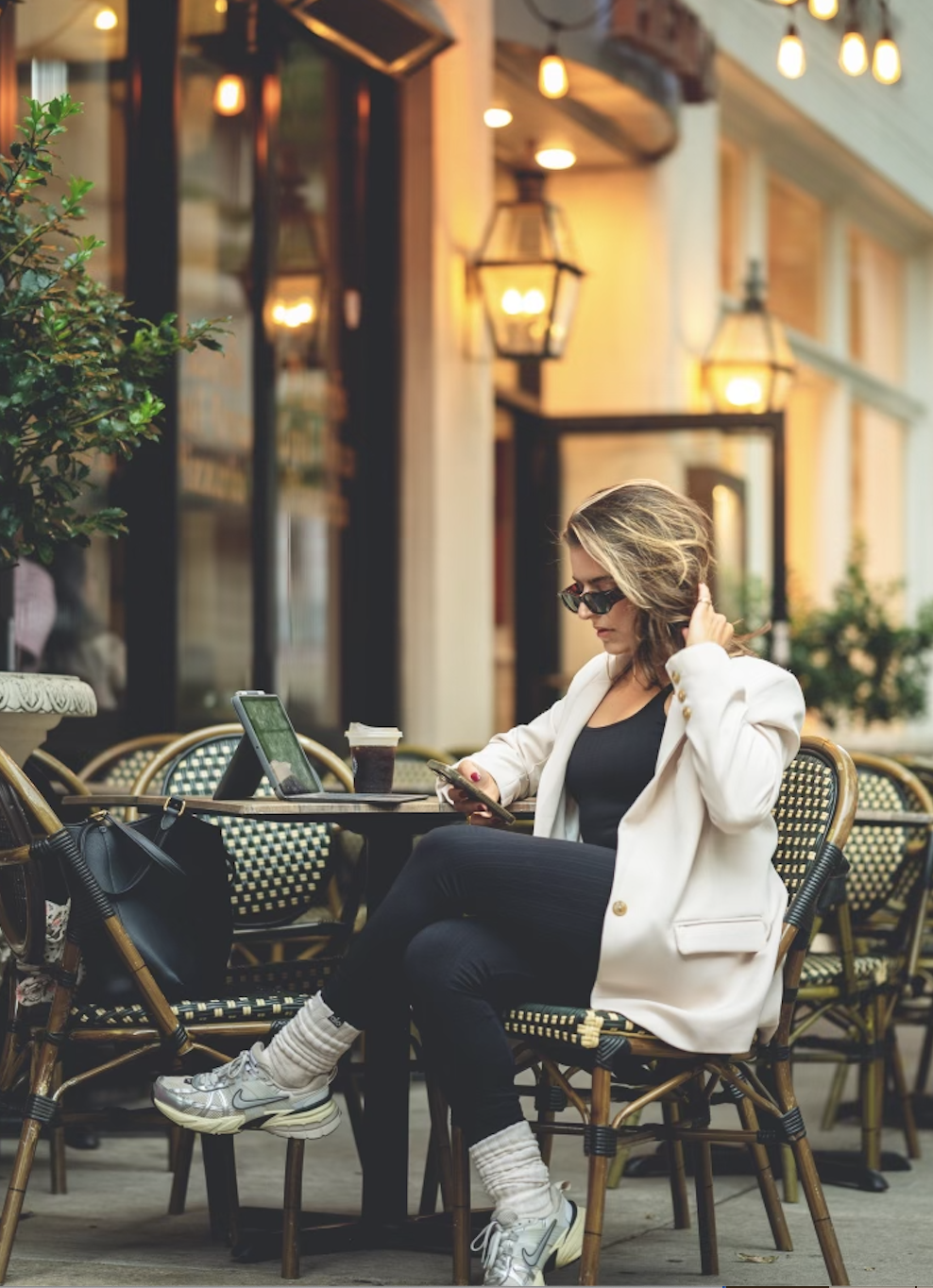 A woman sitting outside at a cafe, looking at her phone, with disposable coffee cups and a tablet on the table, wearing sunglasses, white blazer, black top, black pants, white sneakers, and socks, surrounded by outdoor seating and warm lighting.