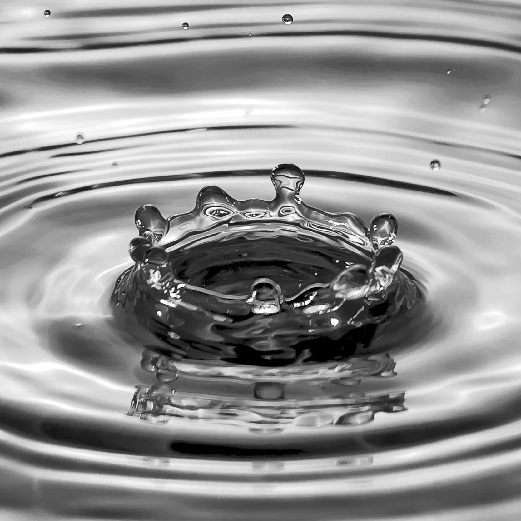 A close-up black and white photograph of a water droplet splash creating a crown-like shape in the water.