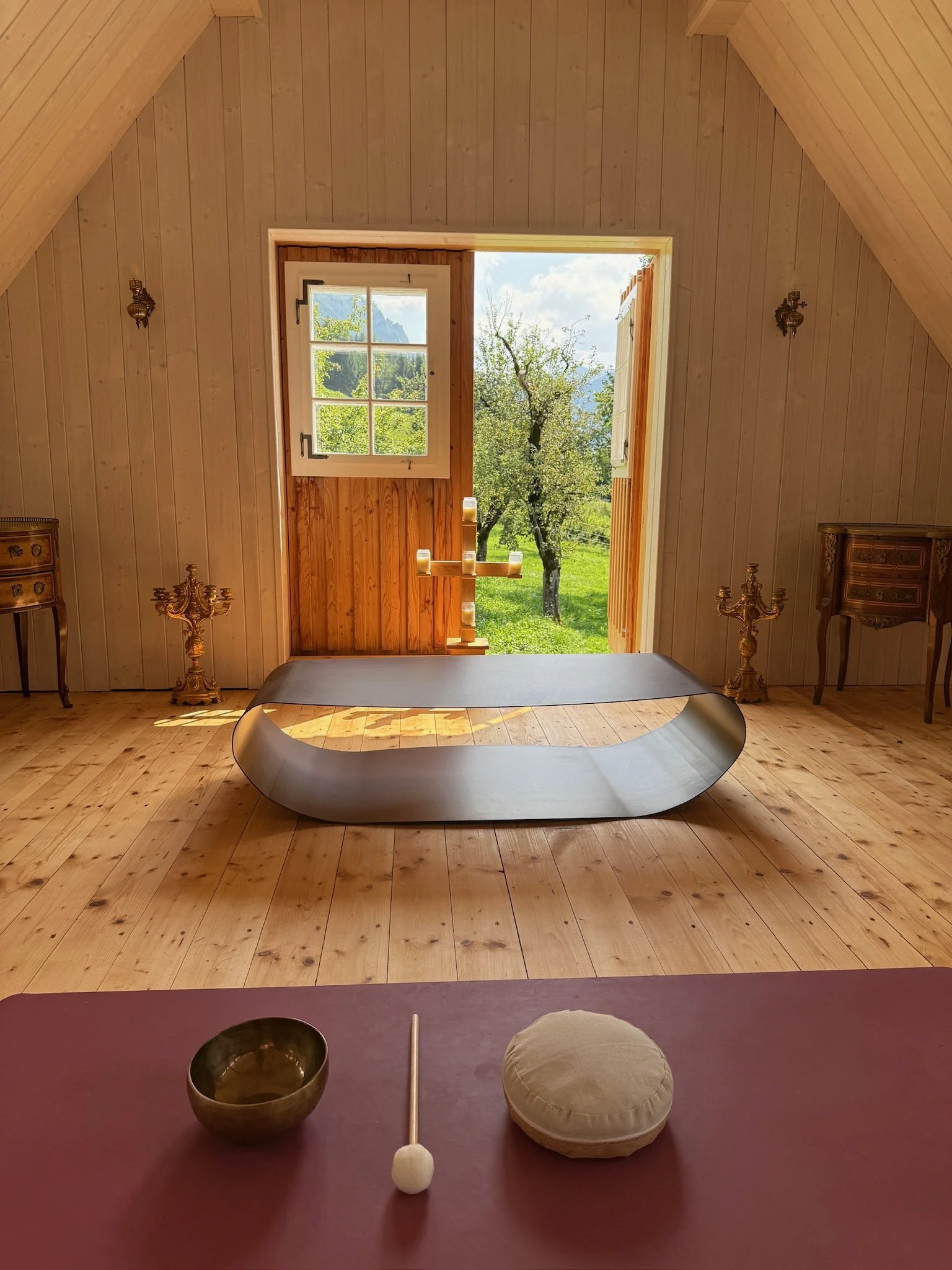 Innenraum eines Holzhauses mit Blick nach draußen auf Bäume und Wiesen, ein modernes silbernes Meditationshallt auf dem Boden, Kerzendocht, Meditationskissen und Schale auf einer roten Oberfläche im Vordergrund.