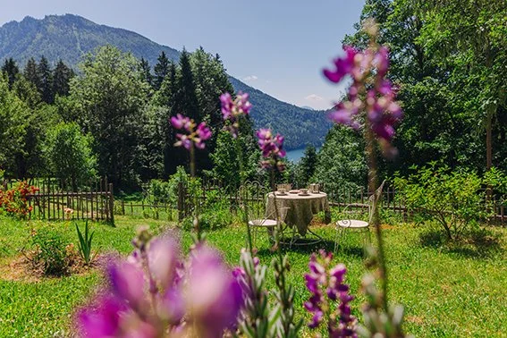 Garten mit Blumen, Tisch und Stühlen vor einem Zaun, mit Wald und Bergen im Hintergrund bei schönem Wetter.
