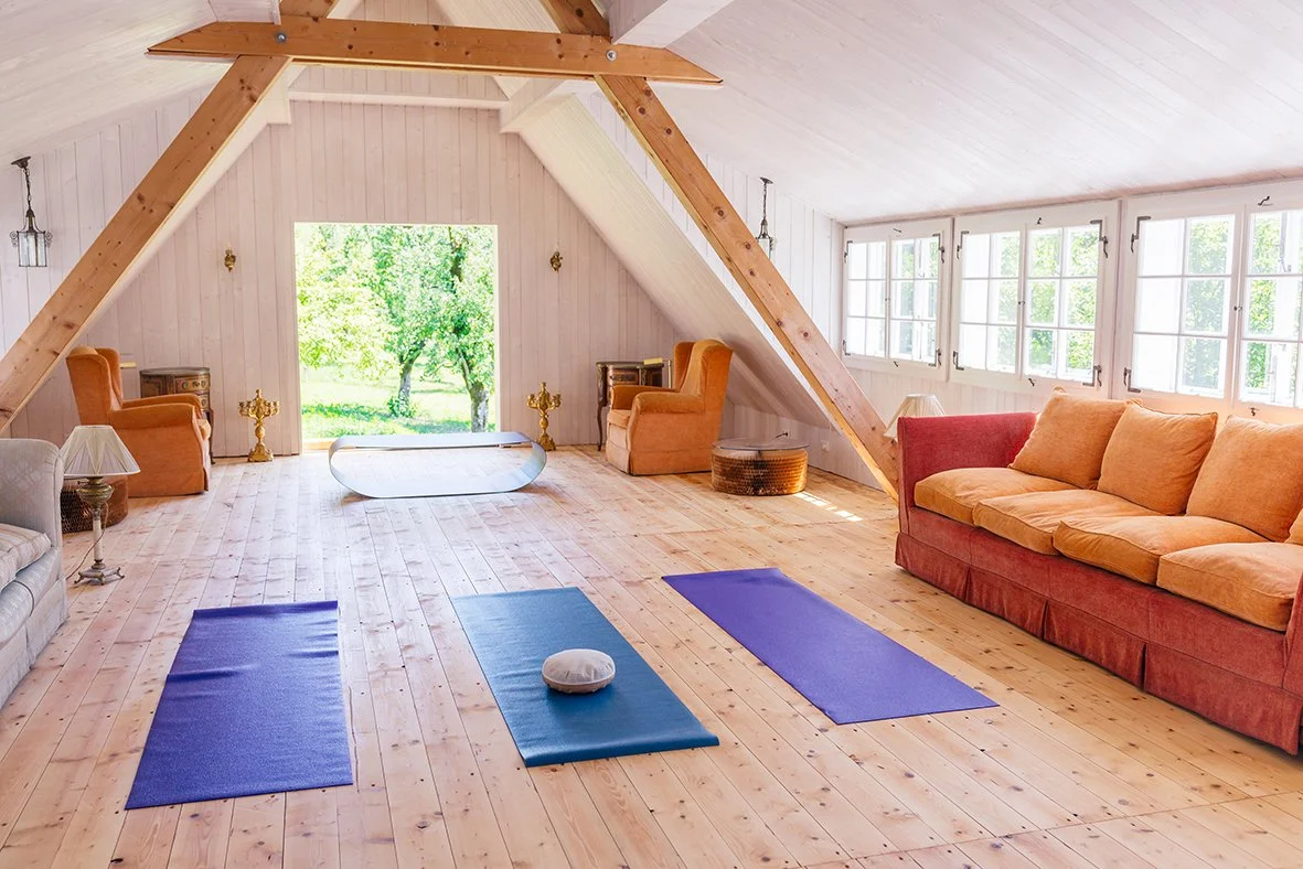 Ein helles Wohnzimmer mit Erdtönen, Holzboden und großen Fenstern. Es gibt Yoga-Matten und Kissen für Meditation oder Yoga, sowie bequeme Sessel und einen Sofa. Der Blick ist nach draußen auf grüne Bäume.
