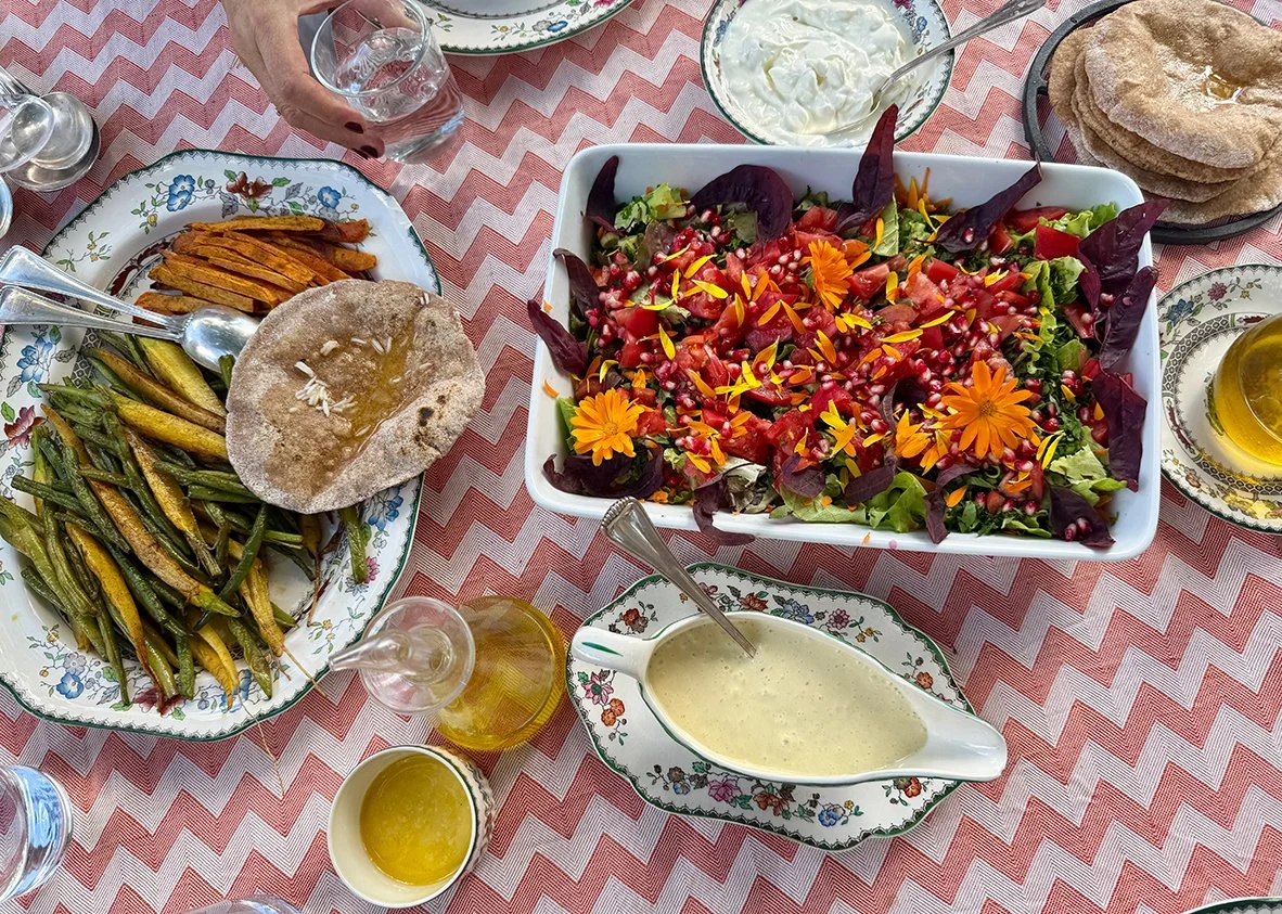 Verschiedene summerische Speisen auf einem rot-weißen Zickzack-Tisch. Es gibt einen großen gemischten Salat mit bunten Blumen und essbaren Dekorationen, eine Platte mit gegrilltem Gemüse, Fladenbrot und eine cremige Sauce. Getränke in Gläsern und kle
