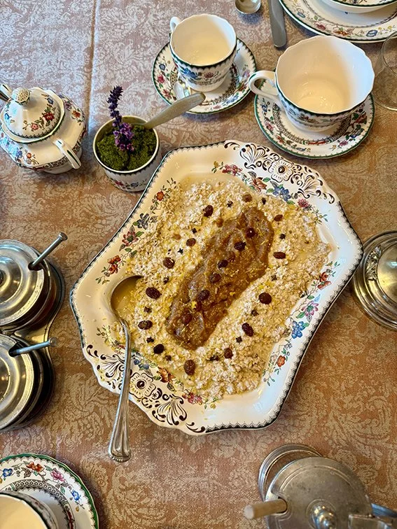 Ein festliches Frühstückstisch mit einer großen Schale Polenta, bedeckt mit Butter und Rosinen, sowie verschiedenen feinen Teegeschirr und einem Topf auf einem gemusterten Tuch.