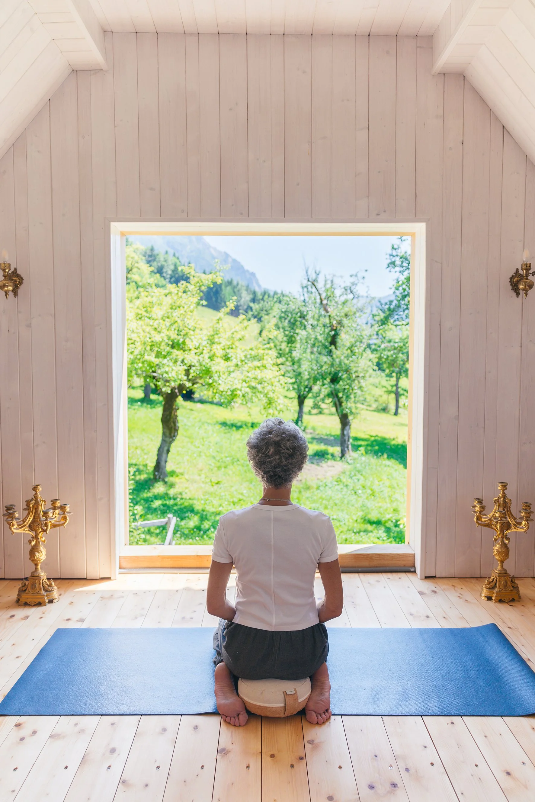 Eine Person im Meditationsraum, die auf dem Boden kniet und nach draußen auf die grüne Landschaft mit Bäumen blickt.