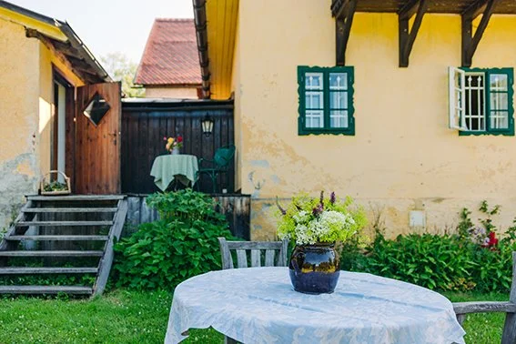 Garten im Hof mit einem runden Esstisch, einer Vase mit Blumen, zwei Stühlen und einem gelben Haus im Hintergrund.