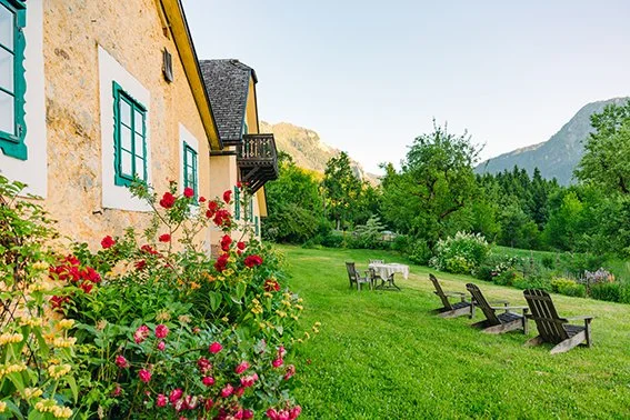 Garten vor einem gelben Haus mit blühenden Rosen, Graskübel, Tischen und Stühlen, umgeben von Bäumen und Bergen im Hintergrund.