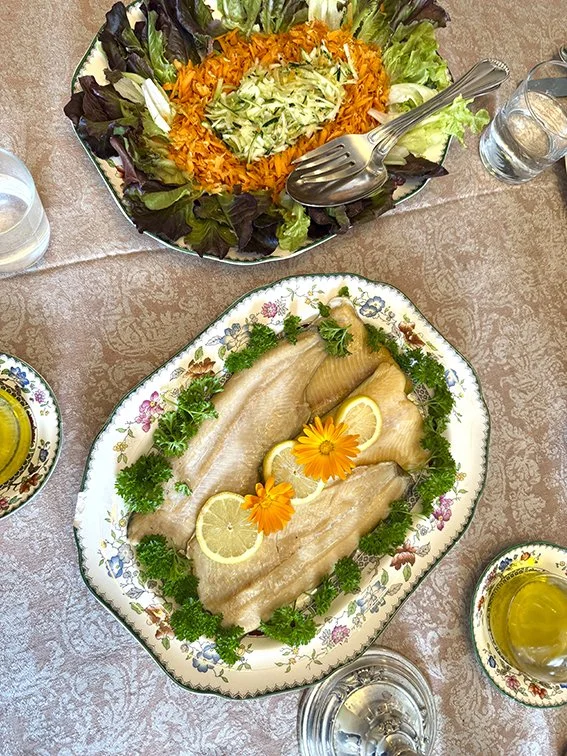 Ein Salat mit buntem Gemüse und ein Gericht mit Fisch, garnierte mit Zitronenscheiben, Blumen und frischen Kräutern auf einem dekorativen Tellern, auf einem Tisch mit Wasser und Olivenöl.
