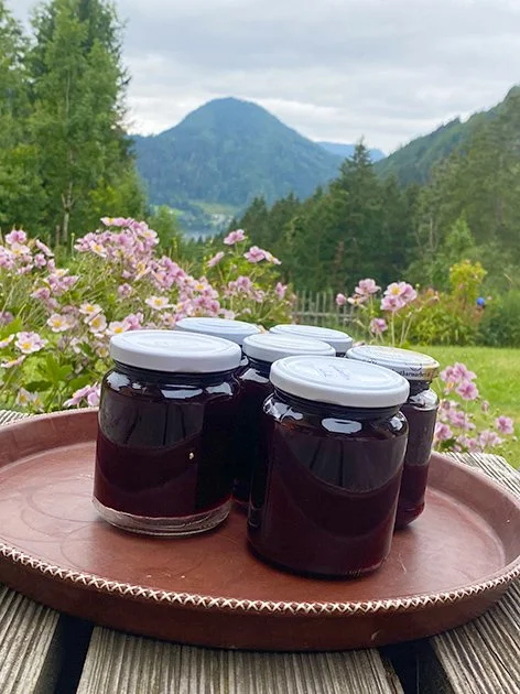 Fünf Gläser mit dunkler Marmelade auf einem großen runden Terrakottateller im Freien, mit pinken Blumen im Vordergrund und Bergen sowie einem bewölkten Himmel im Hintergrund.