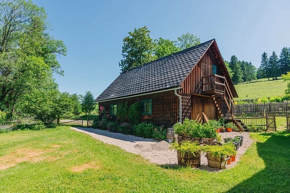Ein traditionelles Holzhaus mit Schornstein in einer grünen, ländlichen Umgebung, umgeben von Bäumen und einem Holzzaun, mit Pflanzen und Blumentöpfen vor dem Haus.