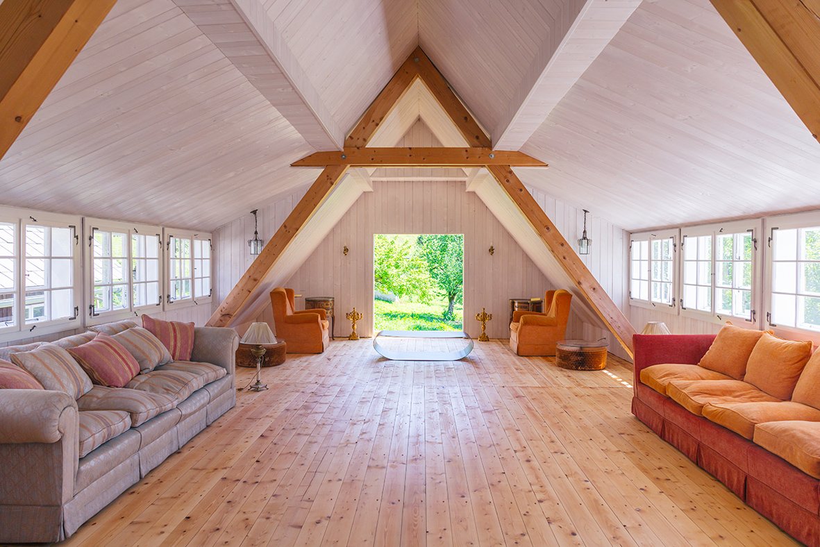 Geräumiger Dachboden mit hellem Holzinterieur, großen Fenstern, bequemen Sofa und Sesseln, Blick auf grüne Landschaft durch offene Tür