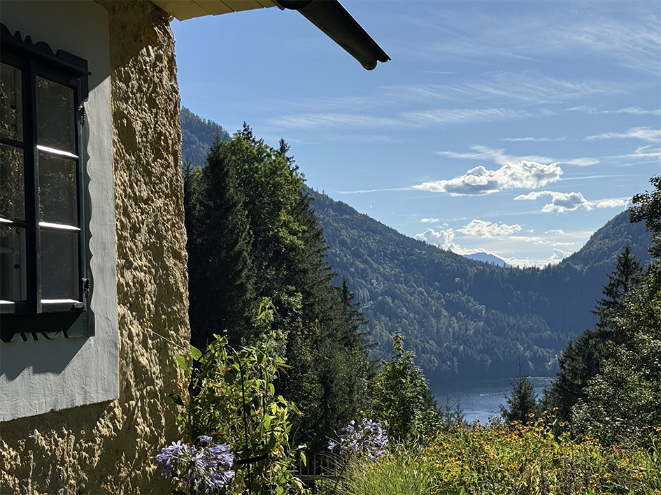 Blick auf einen See, umgeben von bewaldeten Bergen, ein Haus mit einer weißen Wand und einem schwarzen Fensterrahmen im Vordergrund, sonniger Tag mit einigen Wolken am Himmel.