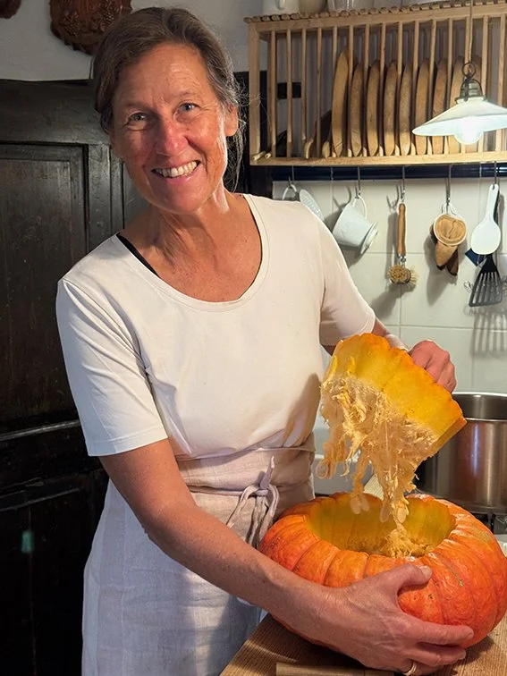 Eine Frau in einer weißen Schürze lächelt und schneidet Kürbis in einer Küche.