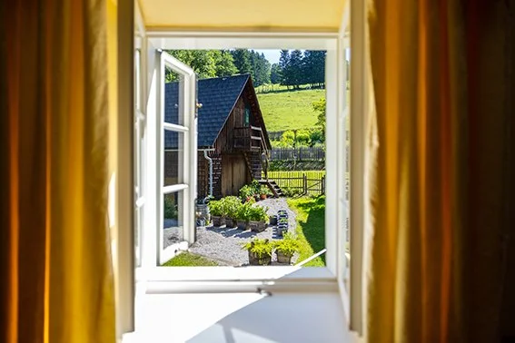 Blick aus einem offenen Fenster auf einen Garten mit einer alten Scheune, grünen Wiesen und Bäumen im Hintergrund.