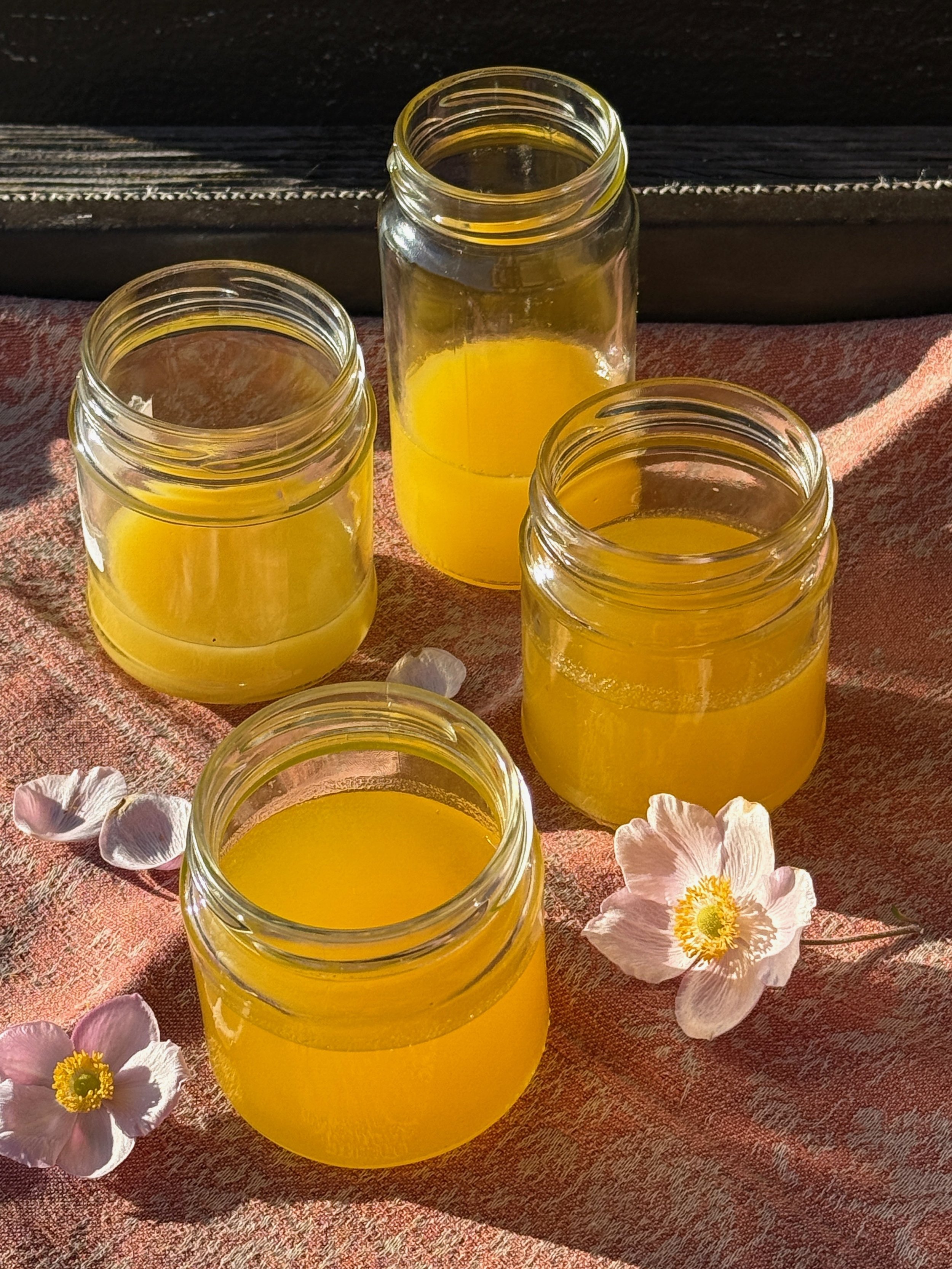 Vier Gläser mit Honig auf einem roten Tuch, umgeben von weißen Blüten, im Sonnenlicht fotografiert.