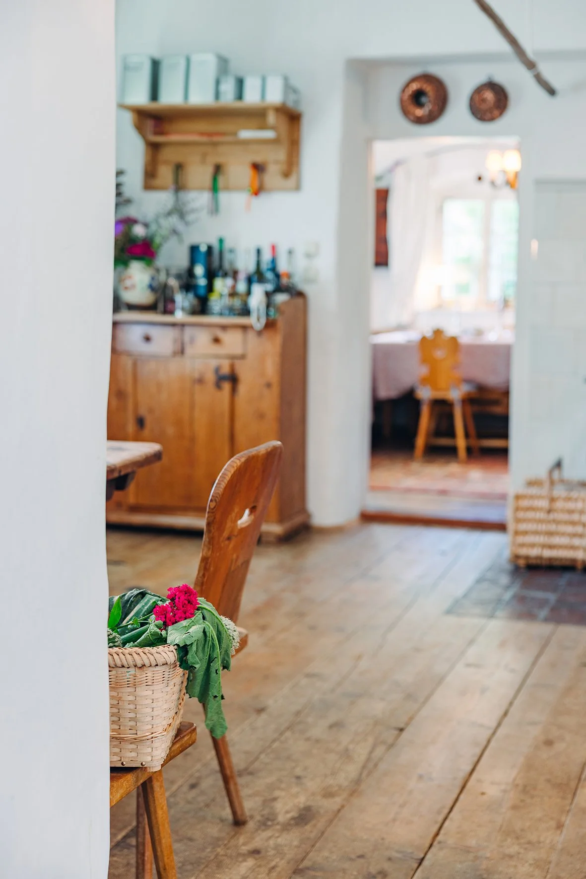 Ein Blick in ein gemütliches, rustikales Wohnzimmer mit Holzböden und Holzmöbeln, im Vordergrund eine Holzstuhllehne mit einem Korb voller pinker Blumen. Im Hintergrund eine Tür, die zu einem weiteren Raum führt, der an einem Fenster mit weißen Vorhä