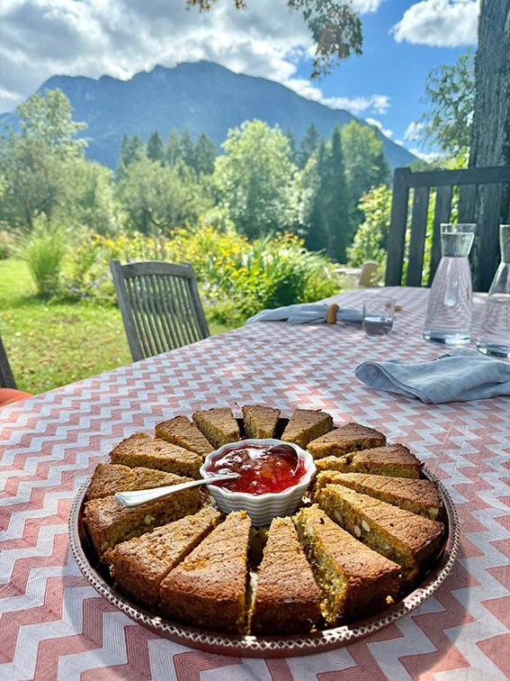 Auf einem Tisch liegt ein Kuchen in Stücke geschnitten, mit einer Portion Erdbeermarmelade in einer kleinen Schüssel in der Mitte, im Hintergrund eine malerische Berg- und Wiesenlandschaft mit Bäumen und Wolken am Himmel.