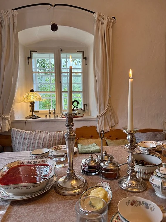 Tisch mit Kerzen, Geschirr und Dekoration in einem rustikalen Raum, mit einem Fenster im Hintergrund, mit weißen Vorhängen und Kerzen auf der Fensterbank.