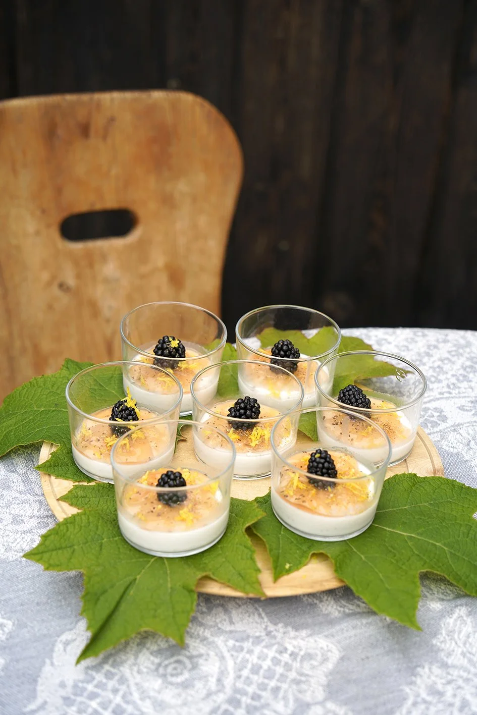 Kleingläser mit Desserts, garniert mit schwarzen Beeren und Zitronenzesten, auf großen grünen Blättern auf einem runden Holzbrett, auf einem weißen Tischdecke mit Blumenmuster, im Hintergrund eine Holzstuhllehne.