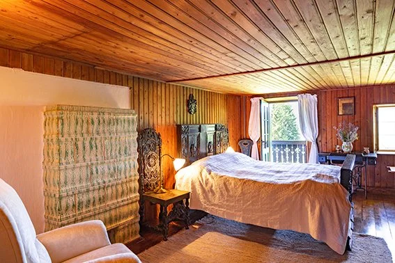 Gemütliches Schlafzimmer mit Holzverkleidung, Doppelbett, Nachttisch, Sessel, Fenster mit Vorhängen und Balkonblick.