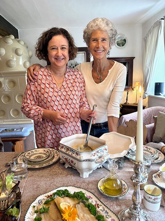 Zwei Frauen, die an einem gedeckten Esstisch in einem Wohnzimmer stehen und lachen. Es gibt Geschirr, eine Kerze, einen Olivenölkrug und Essen auf dem Tisch.