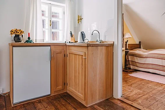 Küchenbereich mit Holzschränken, Wasserkocher, Messerblock, Fenster, neben Schlafzimmer mit Bett im Hintergrund.