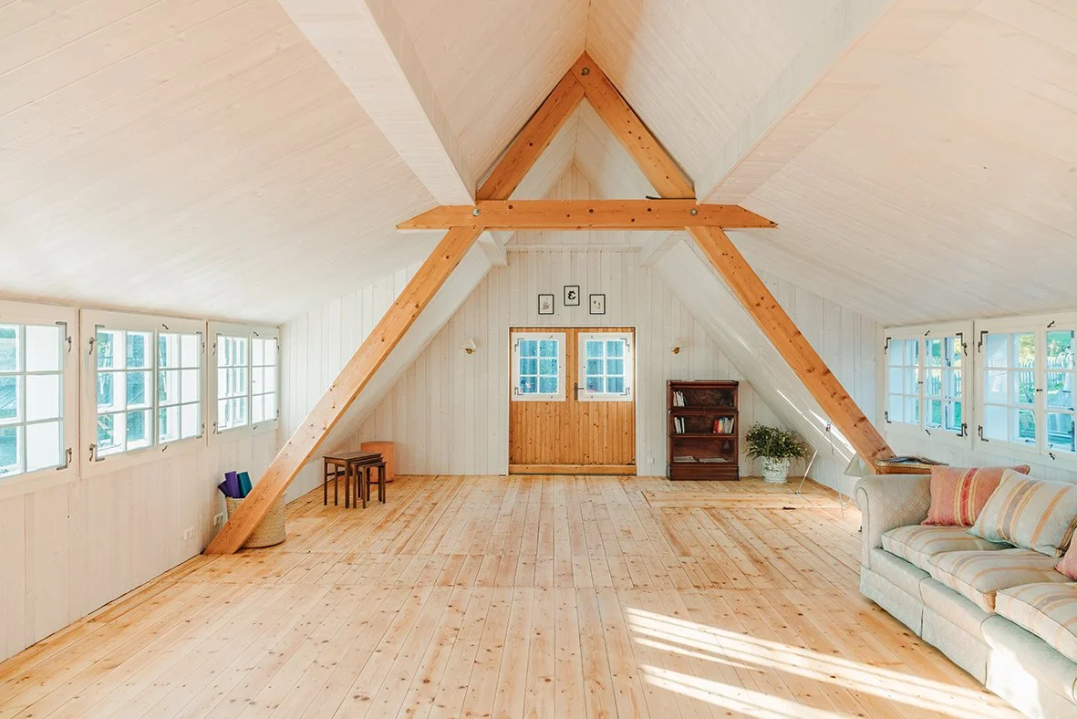 Ein heller, luftiger Dachboden mit Holzboden, weißen Wänden und vielen Fenstern auf beiden Seiten. Es gibt eine Couch mit Kissen, einen kleinen Teppich, Bücherregale, ein paar Stühle und Pflanzen.