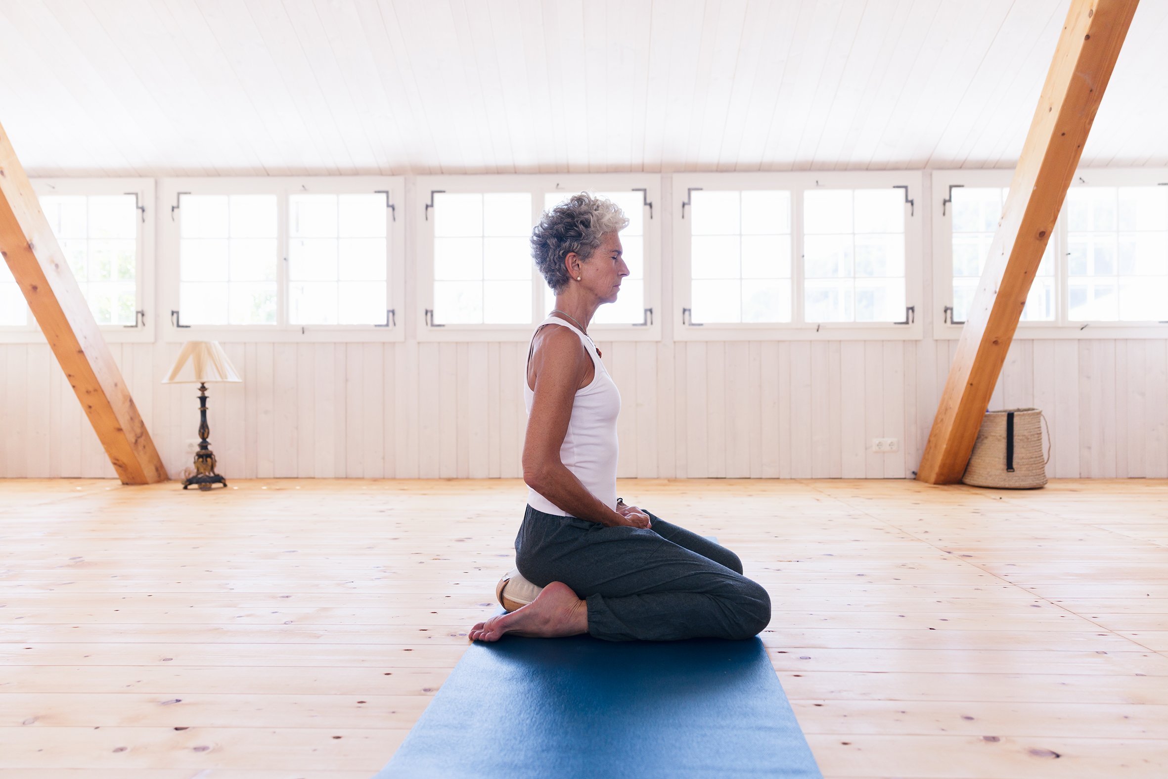 Eine Frau in Yogahose und Top auf Yoga-Matte in einem hellen, Holz-gestalteten Raum in sitzender Yoga-Position