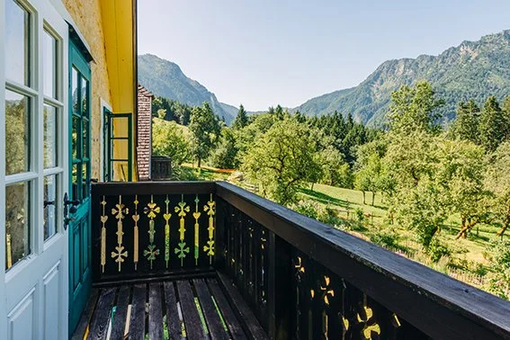 Blick von einem Balkon mit Holzgeländer über grüne Bäume und eine bergige Landschaft bei sonnigem Wetter.