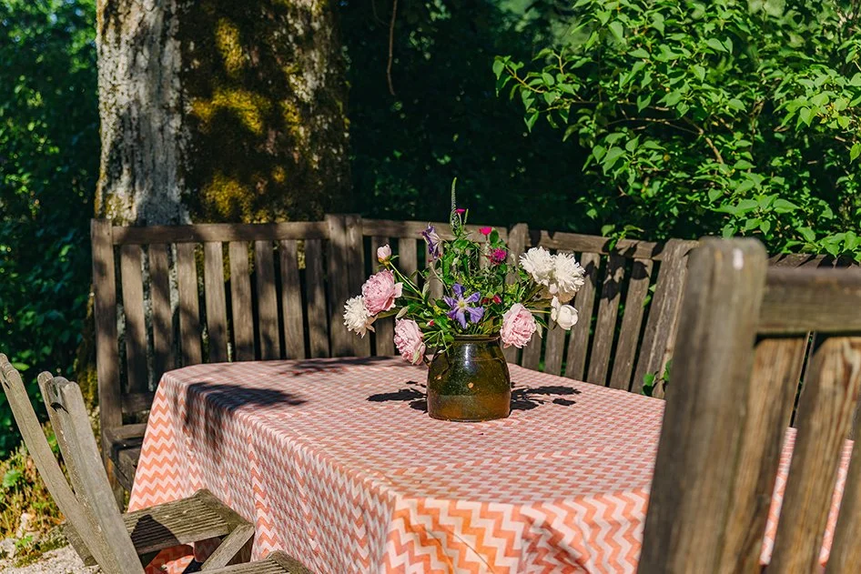 Ein Tisch mit einem Zickzack-Muster-Tuch, in der Mitte eine Vase mit bunten Blumen, im Freien mit Bäumen im Hintergrund.