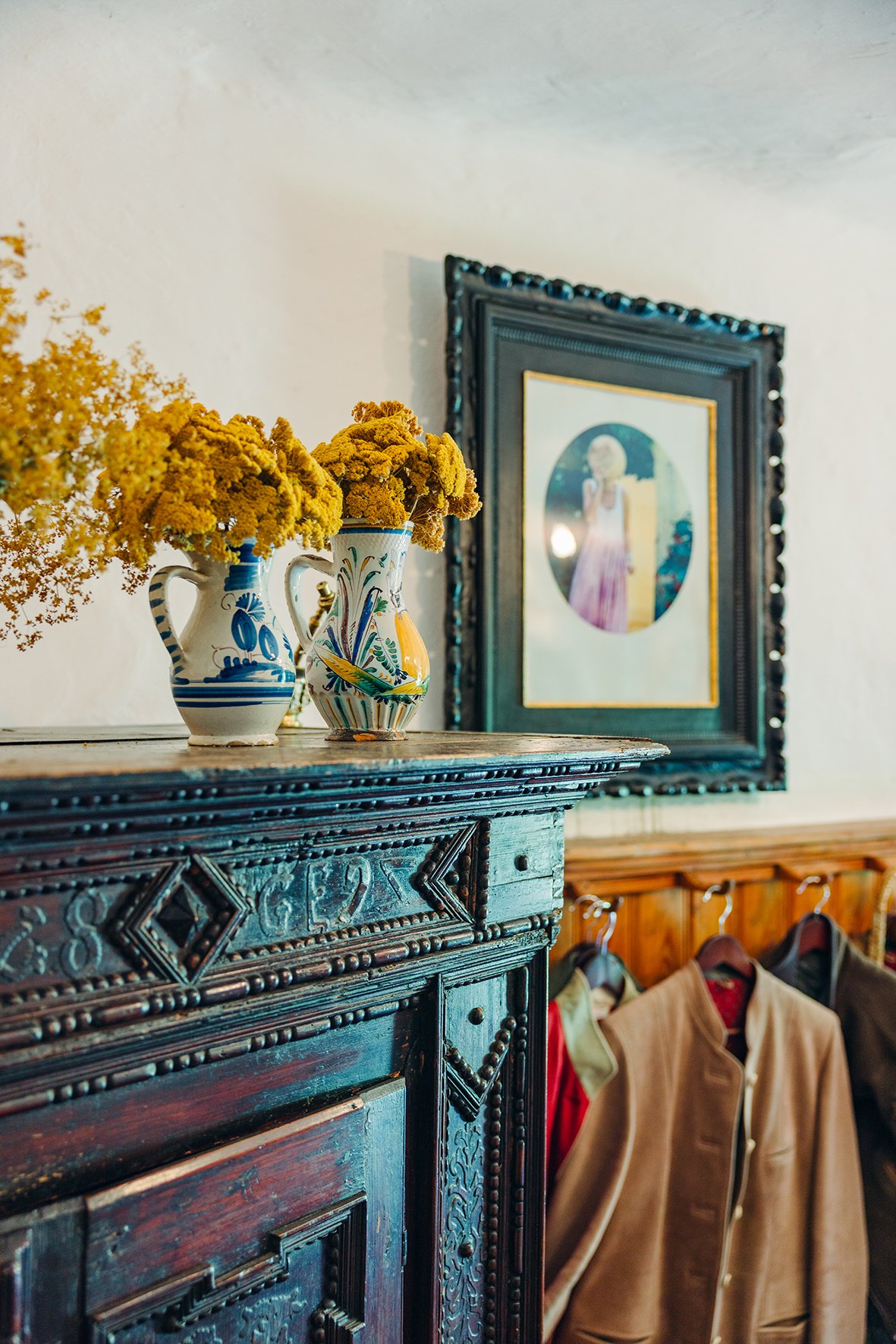 Vasen mit gelben Blumen auf einer Holzkommode, im Hintergrund ein gerahmtes Gemälde an der Wand, darunter Jacken an Kleiderhaken.