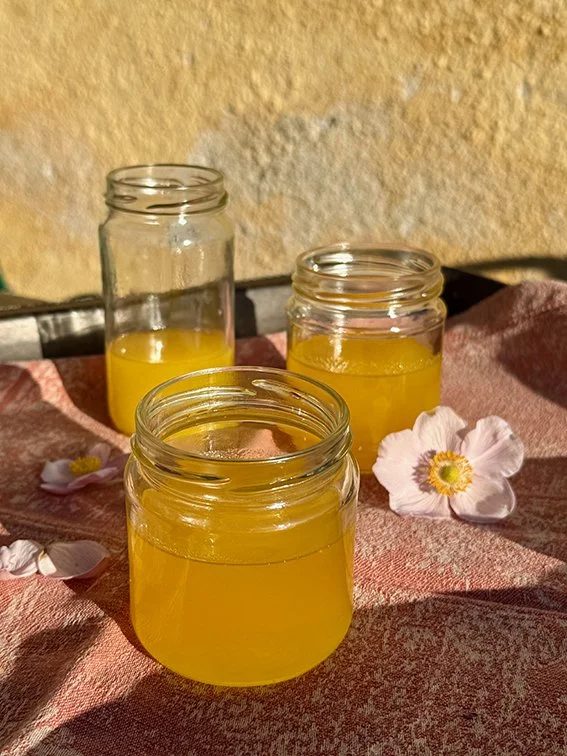 Drei offene Gläser mit gelber Flüssigkeit stehen auf einer rosa Stoffdecke, umgeben von weißen Blumenblüten, im Sonnenlicht neben einer gelben Wand.