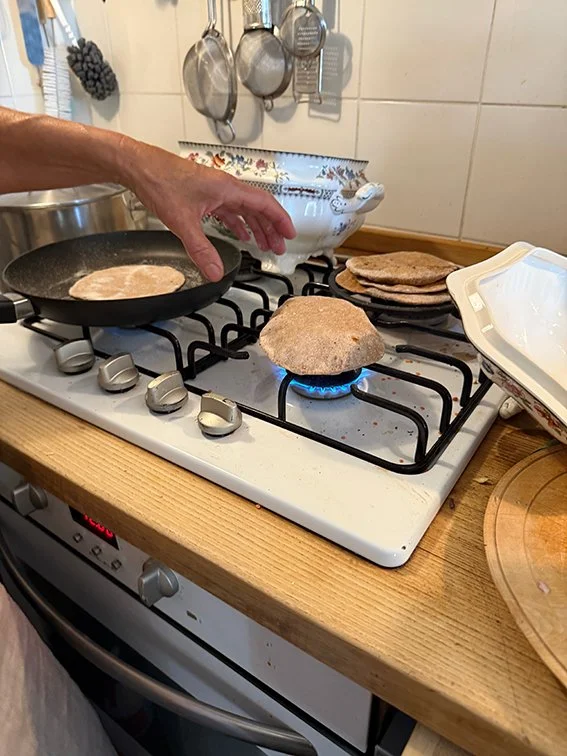 Person kocht auf einem Küchenherd, mit Pfanne, in der eine Teigflade brät, und Fladenbrot, das auf einem Backblech liegt.
