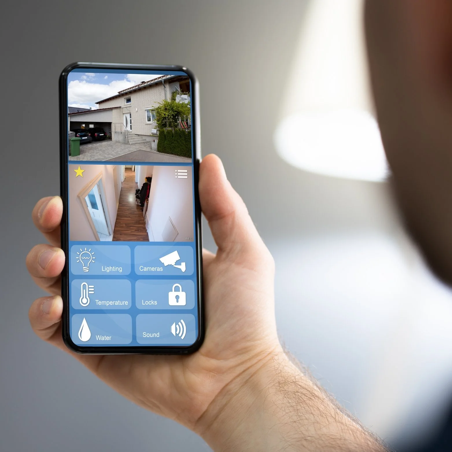 A person holding a smartphone controlling smart home features such as lighting, cameras, temperature, locks, water, and sound, with interior and exterior views of a house on the screen.